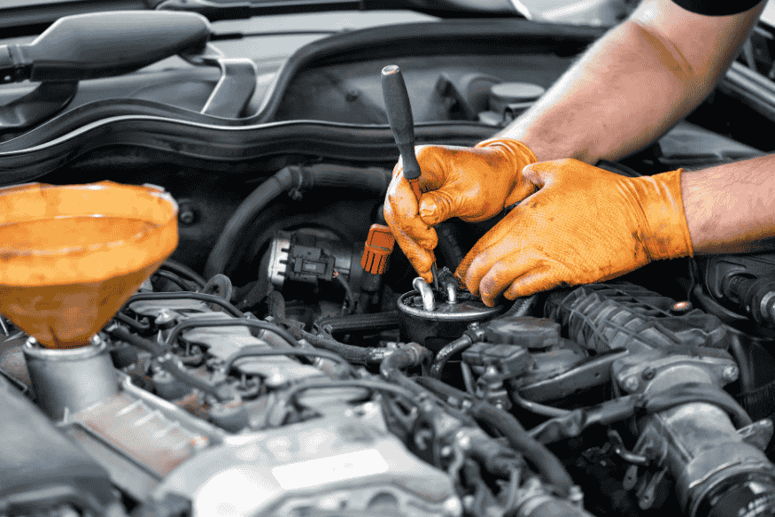 Mãos de mecânico usando luvas laranja trocando componente no motor de um carro com funil de óleo.